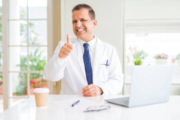 Fototapeta na wymiar Middle age doctor man wearing white medical coat working with laptop at the clinic doing happy thumbs up gesture with hand. Approving expression looking at the camera with showing success.