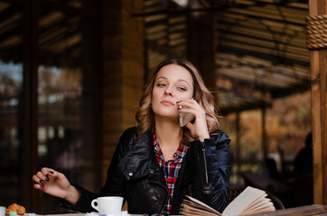 Attractive young woman calling with cell telephone while sitting alone at the table with book in street cafe, drinking coffee or tea.  Relaxing in cafe during free time