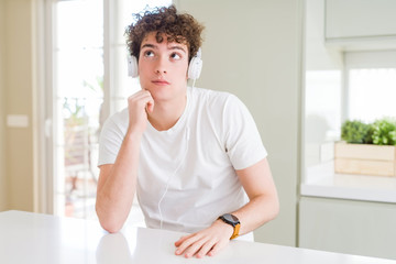 Fototapeta premium Young man listening to music wearing headphones at homes with hand on chin thinking about question, pensive expression. Smiling with thoughtful face. Doubt concept.