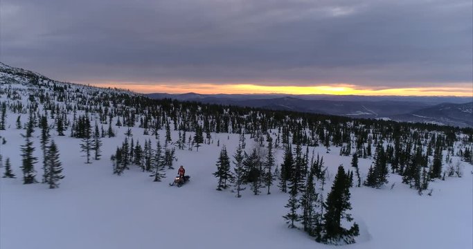 a guy on a mountain snowmobile rides on the background of a beautiful sunrise in a forest mountain valley with fields of snow. snowmobile traffic in the mountains. stock video footage. Aerial shot