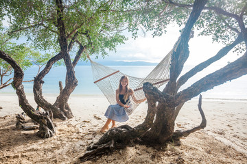 Young happy girl sitting in the hammock between green trees on the tropical beach near the sea