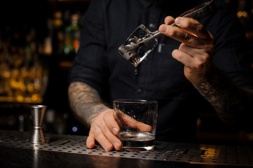 Bartender adding to an alcoholic cocktail in the glass a big ice cube
