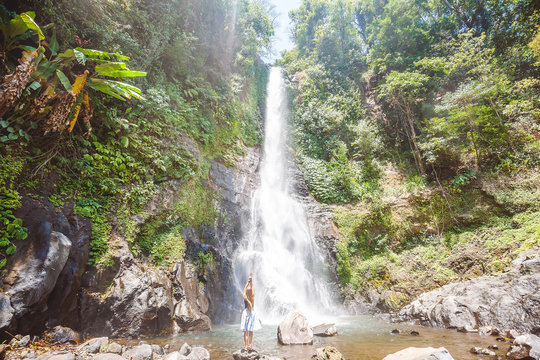 Young woman with long hair dressed in skirt standing back on the stones in front of Gitgit waterfall among green tropical trees on the north of Bali island, Indonesia
