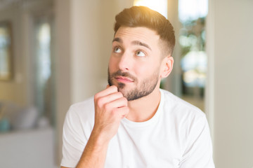Young handsome man wearing casual white t-shirt at home with hand on chin thinking about question, pensive expression. Smiling with thoughtful face. Doubt concept.