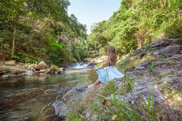 Young woman with long hair sitting back on the rocks near small river with clean water among green tropical trees and plants while sky is blue on the north of Bali island, Indonesia