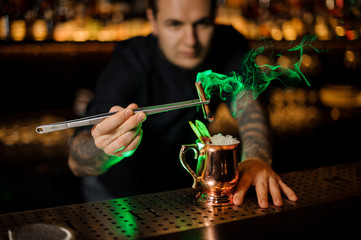 Bartender adding to the cocktail with dried orange a smoked cinnamon with tweezers in the red light