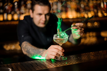 Bartender adding to a cocktail glass with dried orange a smoked cinnamon with tweezers