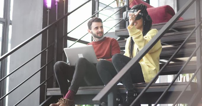 In a modern business building one guy and African lady spending a good time together sitting on the modern stairs lady trying for first time the VR she touching virtual with finger to select favorite