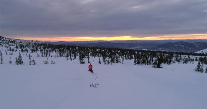 snowmobile rides to the left of the camera, looks at the frame and quickly rides a snowmobile in a beautiful mountain valley at sunrise. drone's coming.  stock video footage. Aerial drone shot