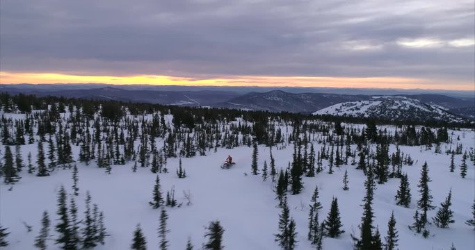 a guy on a mountain snowmobile rides on the background of a beautiful sunrise in a forest mountain valley with large fields of snow. movement on a snowmobile in the mountains, the camera flies behind 