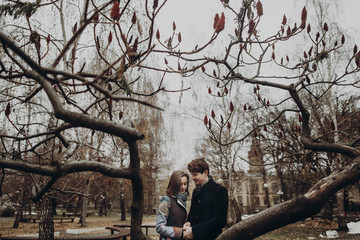 Fototapeta premium romantic stylish couple hugging gently, holding hands in autumn park under unusual tree. man and woman embracing. family togetherness concept. calm atmospheric moment