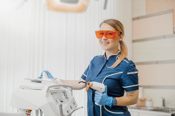 Dentist in blue uniform and orange glasses standing next to dental equipment.