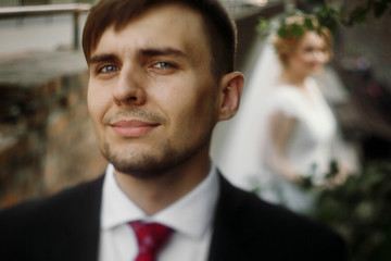 Happy, handsome groom with a beard in stylish suit smiling outdoors, with bride standing in the background, newlywed man portrait, face closeup