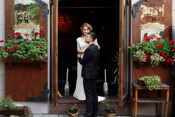 Beautiful romantic newlywed couple hugging near flower bouquets at luxury french restaurant outdoors, emotional blonde bride being embraced by handsome groom portrait