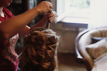 Professional stylist doing hairstyle for beautiful woman, blonde bride getting her hair done by hairdresser, morning wedding preparation closeup