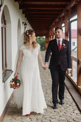 Happy bride holding hands with groom, beautiful blone bride in white wedding dress walking with handsome groom on paving road near wall, happy newlyweds