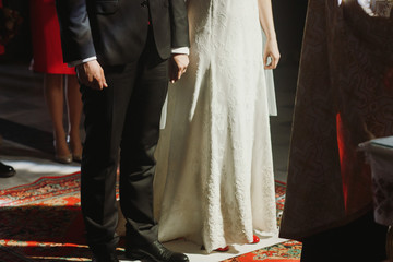 Spiritual couple, handsome groom in stylish suit and bride in vintage white dress with bouquet standing in church during wedding ceremony, hands closeup