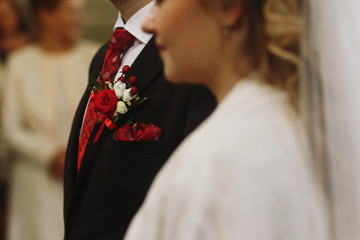 Closeup of red rose boutonniere on groom's suit during wedding ceremony in church, red flower...