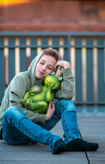 a young boy sits on a terrace on a wooden floor and hugs a large green frog in a golden crown
