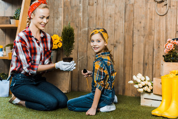 attractive woman holding green plant and sitting with cheerful daughter on grass