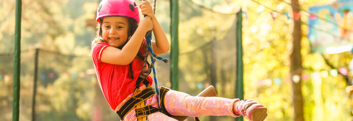 Little beautiful girl climbs on rope harness in summer city park.