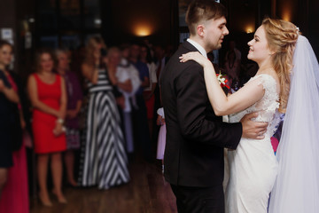 Romantic bride and groom dancing and holding hands at wedding reception in restaurante, newlywed couple first dance at evening party, guests looking in the background