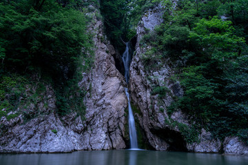 A high waterfall falls from a cliff into a clear lake in the evening.