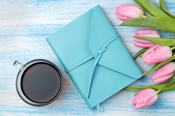 Beautiful bouquet of flowers of pink tulips and a blue notebook and a cup of coffee on a blue wooden background. top view. Spring. holidays.
