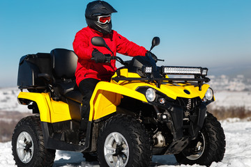 Photo of attractive young man in red warm winter clothes and black helmet on the ATV 4wd quad bike stand in heavy snow with deep wheel track. Moto winter sports.