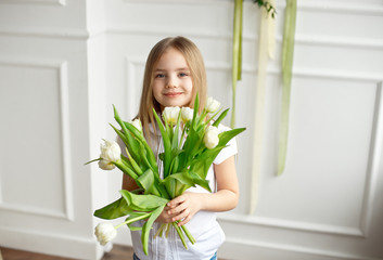 Portrait of a very cute pretty blonde girl in a white shirt against a white wall with a bouquet of white tulips