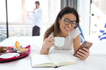 Beautiful smiling woman using her mobile phone and drinking coffee while lying on bed in hotel room.