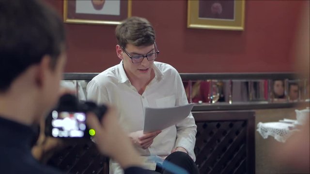 The actor, sitting in front of the audience in a cozy interior, reads prose from paper