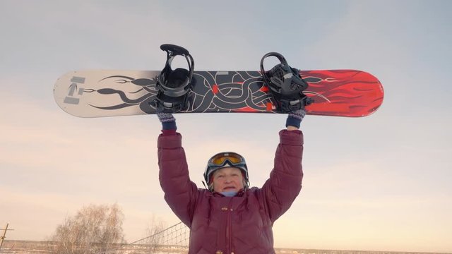 Cheerful older woman rising up snowboard on snowy slope at winter resort. Happy senior woman snowboarding rejoicing new season on snowy resort. Winter sport concept golden and glorious senior woman