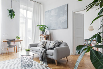Stylish scandinavian living room with design furniture, plants, bamboo bookstand and wooden desk. Brown wooden parquet. Abstract painting on the white wall. Nice apartment. Modern decor of bright room