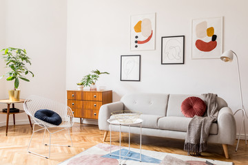 Stylish vintage decor in a spacious flat interior with design grey sofa, armchair, retro commode and posters on the wall. Brwon wooden parquet, stylish carpet and plants. Bright  living room.