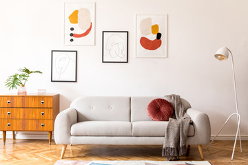 Stylish vintage decor in a spacious flat interior with design grey sofa, armchair, retro commode and posters on the wall. Brwon wooden parquet, stylish carpet and plants. Bright  living room.