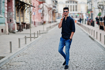 Stylish indian model man in casual clothes, black shirt and sunglasses posed outdoor at street of India and speaking at telephone.