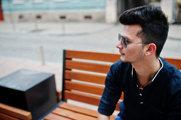 Stylish indian model man in casual clothes, black shirt and sunglasses posed outdoor at street of India and sitting on bench.