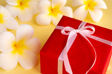 Red gift box with tropical white flowers (plumeria) on yellow background. Concept for greeting...
