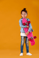 Stylish little child girl with skateboard in jeans clothes on orange background