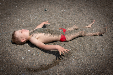 The boy lies on the beach and is covered with small pebbles.