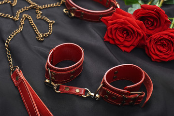 Erotic games. Top view of BDSM leather stuff: red handcuffs and roses against of black silk fabric