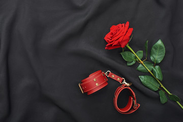 BDSM fantasy play. Top view of red leather handcuffs and rose against of black silk fabric