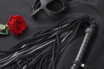 Role playing. Top view of bdsm leather kit ( black whip and mask) with red rose on black silk