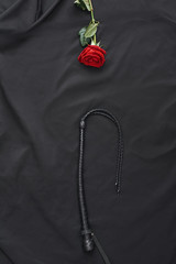 Enter the world of BDSM. Top view of black leather whip and red rose against of black silk
