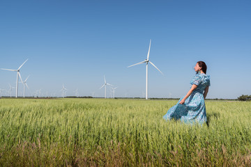 Freedom concept. Beautiful woman in blue dress on green field with wind generator turbines and blue sky background 