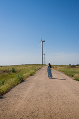 Beautiful woman with long hair in a blue dress walks on road between green fields and wind turbines on blue sky background