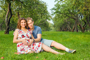 A man in a blue t-shirt and denim shorts sitting with a pregnant girl in a beautiful dress on the green grass on a summer day against the Apple orchard