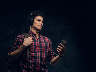 Pensive young guy in wireless headphones listens to music and keeps the smartphone in a studio against the background of the dark wall