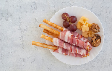 Breadsticks grissini with prosciutto or bacon with slices of lambere cheese, grapes and walnuts on a white plate, top view, close-up. Copy space.